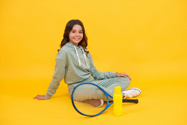 veselé dítě sedět ve sportovním oblečení s squash raketa a láhev vody na žlutém pozadí, zdraví. - Fotografie, Obrázek