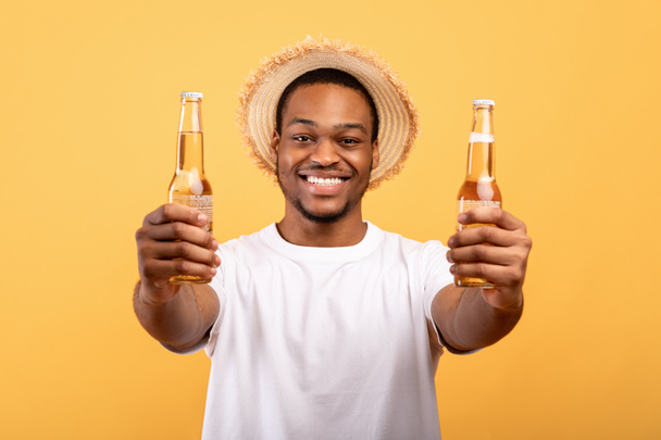 Attractive black guy in t-shirt and straw hat holding two bottles of beer in outstretched hands on yellow background - Photo, Image