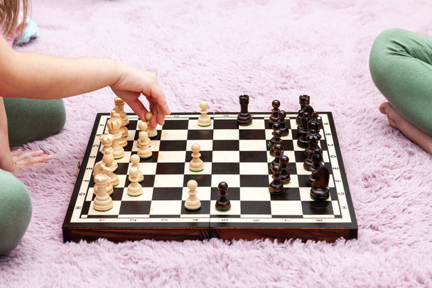Двоє анонімних невідомих дітей шкільного віку, маленькі дівчата грають у шахи вдома, шахова дошка лежить на килимі на землі, одна дитина робить крок, рухаючи пішак
 - Фото, зображення