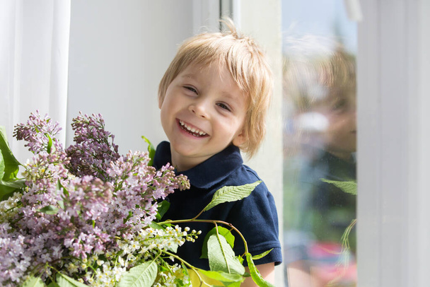 Close portrait of a beautiful blond child, toddler boy with vase with lilacs at home, sitting on the window - Φωτογραφία, εικόνα
