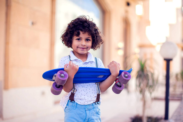 Kleine jongen met krullend haar met skateboard outdoor. Ontwerp voor kinderen en amusement - Foto, afbeelding