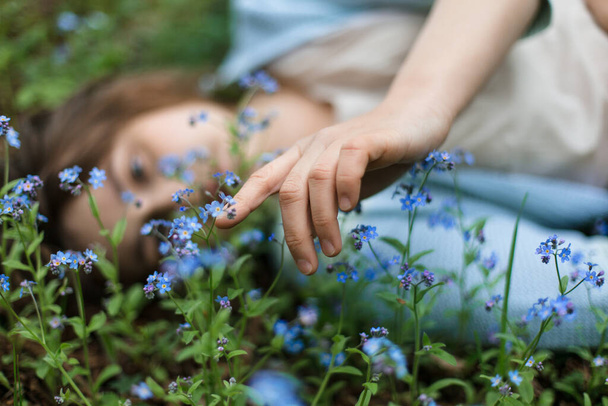 Girl lies in meadow in flowers of forget-me-nots and runs her hand over them. - Foto, Imagen