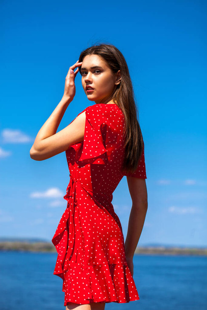 Portrait of a young beautiful girl in a short red dress posing against the blue sky - Фото, зображення