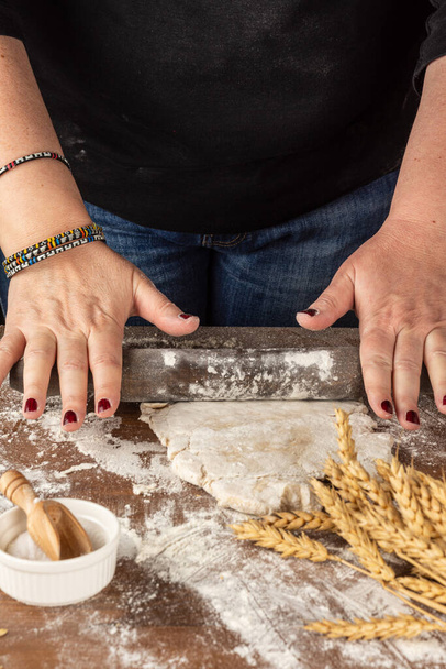 Vista superior de las manos de la mujer amasando masa de pizza con rodillo, sobre mesa de madera con harina, sal y trigo, enfoque selectivo, fondo negro, vertical - Foto, Imagen