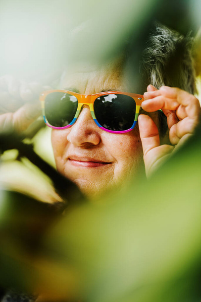 Portret van lesbische oma die een zonnebril draagt met de gay pride vlag. Oude dame tussen groene planten. Collectief overleg. - Foto, afbeelding