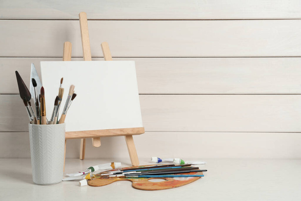 Easel with blank canvas, brushes, paints and palette on white wooden table. Space for text - 写真・画像
