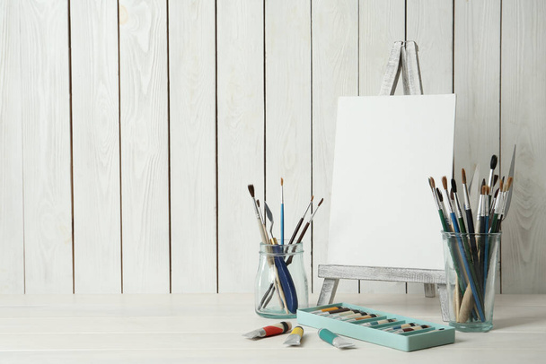Easel with blank canvas, paints and brushes on white wooden table. Space for text - 写真・画像