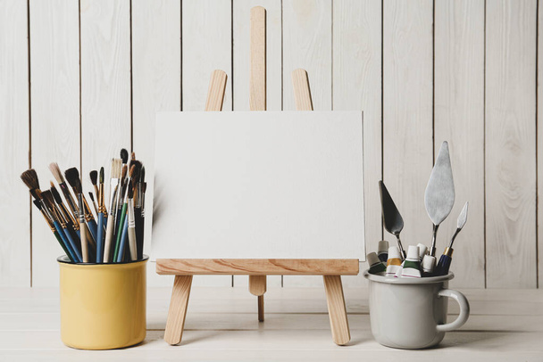 Easel with blank canvas, paints and brushes on white wooden table - Foto, Bild