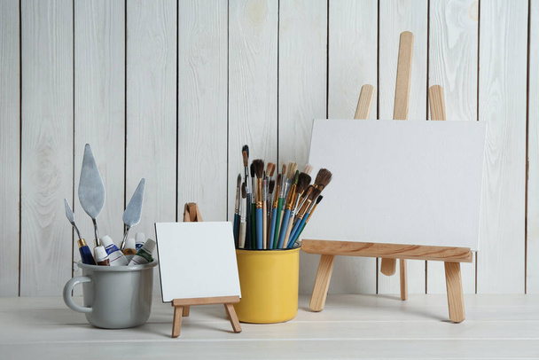 Easels with blank canvases, paints and brushes on white wooden table - Фото, изображение