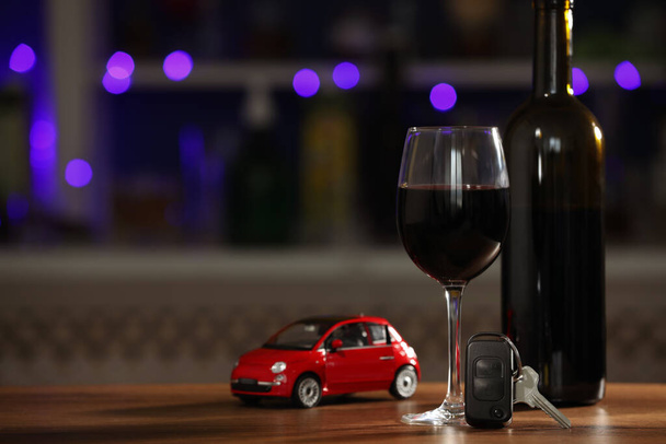 Car key, glass of alcohol near toy auto on wooden table against blurred lights, space for text. Dangerous drinking and driving - Photo, Image
