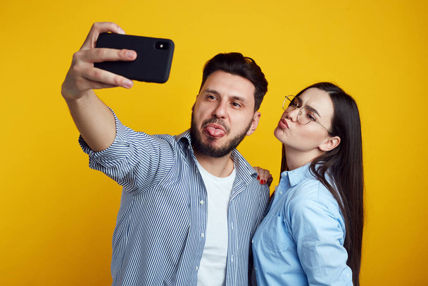 Забавная пара делает гримасу, делает селфи на смартфоне над желтой стеной - Фото, изображение