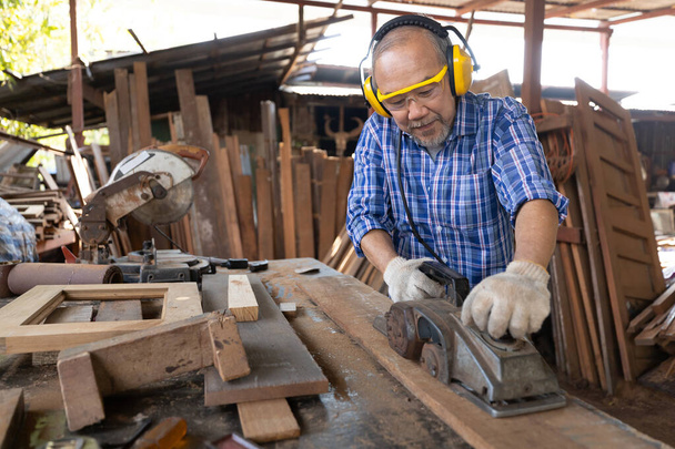 Kıdemli Asyalı marangoz gözlük ve kulaklık takıyor, atölyedeki bir tahta parçasında elektrikli odun planörleri kullanıyor. - Fotoğraf, Görsel