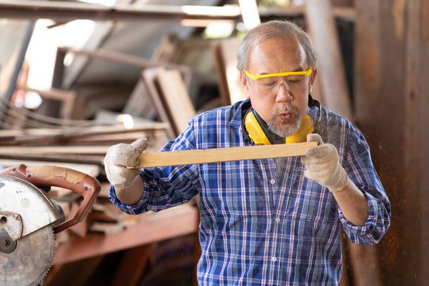 παλαιός ασιάτης ξυλουργός που πετάει πριονίδια από σανίδα ξύλου σε εργαστήριο ξυλουργικής - Φωτογραφία, εικόνα