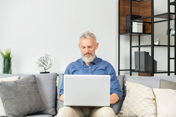 Moderner bärtiger Mann mittleren Alters sitzt mit Laptop zu Hause auf der Couch - Foto, Bild