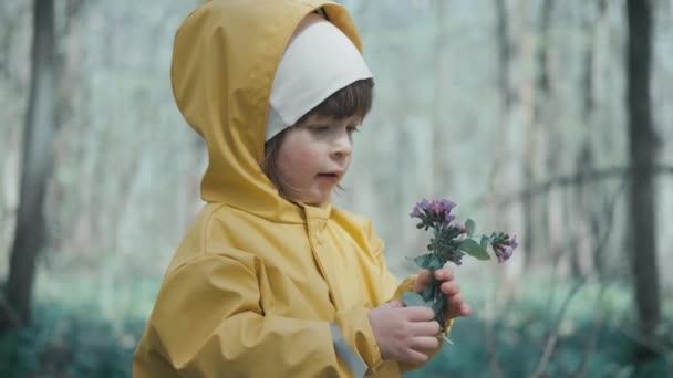 Child in yellow raincoat with hood stands in forest and holds spring flowers - Footage, Video