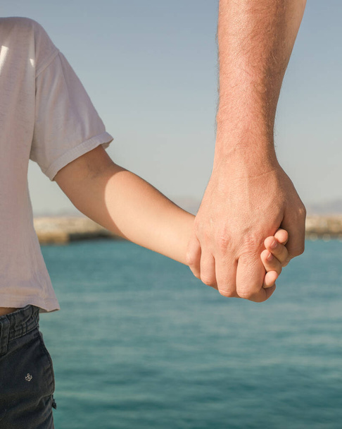 Pieni poika pitelee isäänsä kädestä. Nuori mies, pieni poika kävelemässä rannalla, isipäivä. Onnellinen lapsuus isän kanssa. - Valokuva, kuva