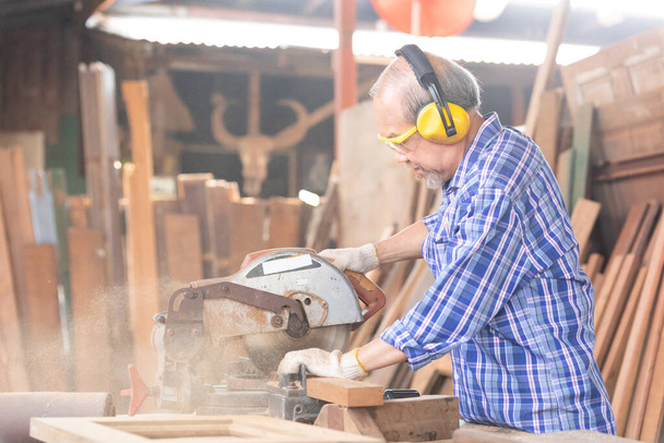 Kıdemli Asyalı marangoz gözlük ve kulaklık takıyor, elektrikli testere kullanarak atölyedeki bir tahta parçasında tahta kesiyor. - Fotoğraf, Görsel