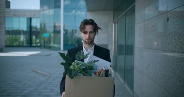 Outdoor portrait of depressed fired male employer walking out of office building with box of personal stuff - Footage, Video