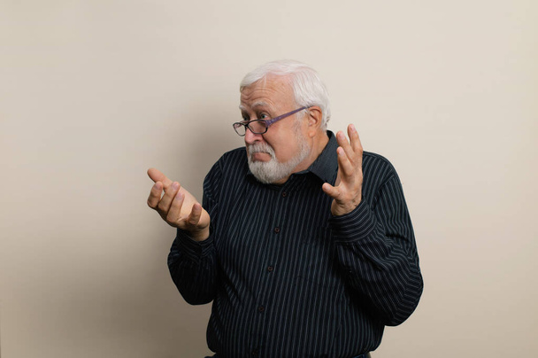 anciano hombre de pelo gris con una barba en gafas y una camisa negra sorprendentemente extiende sus brazos, emociones en el marco, retrato de cintura, fondo claro, estudio - Foto, imagen