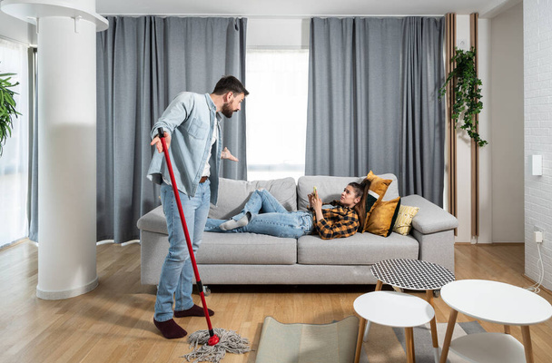 Man wipes and cleans the floor of an apartment with mop and argues with a lazy woman lying on a sofa and spends time on social media on a smartphone end dont want to help him in housework - Photo, image