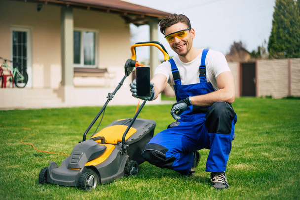 Young man mows the lawn using an electric lawn mower in a special worker suit near a large country house in the backyard - Valokuva, kuva