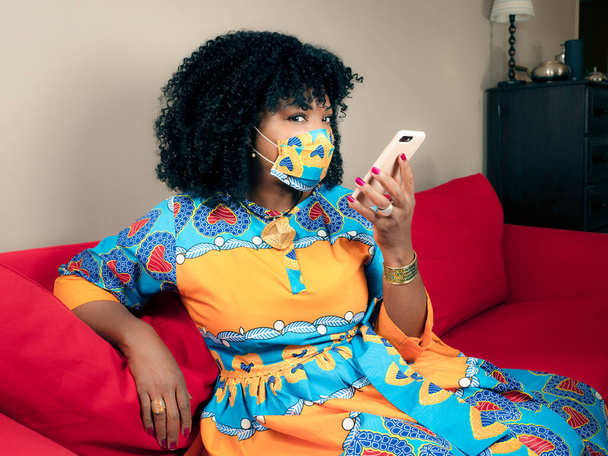 Maske takan ve elinde telefon tutan Afro-Amerikalı bir kadın kırmızı bir kanepede oturuyor. - Fotoğraf, Görsel