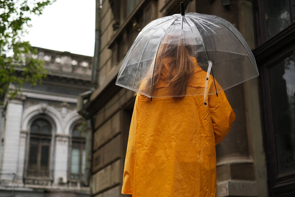 όμορφη γυναίκα σε ένα κίτρινο αδιάβροχο βόλτες γύρω από την πόλη με μια διάφανη ομπρέλα στη βροχή. - Φωτογραφία, εικόνα