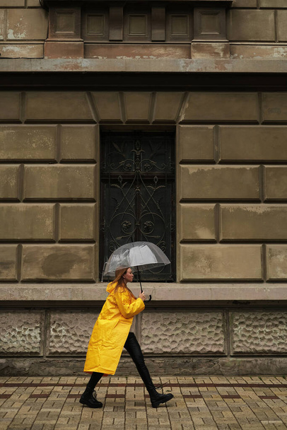 Schöne Frau in gelbem Regenmantel geht mit durchsichtigem Regenschirm im Regen durch die Stadt. - Foto, Bild