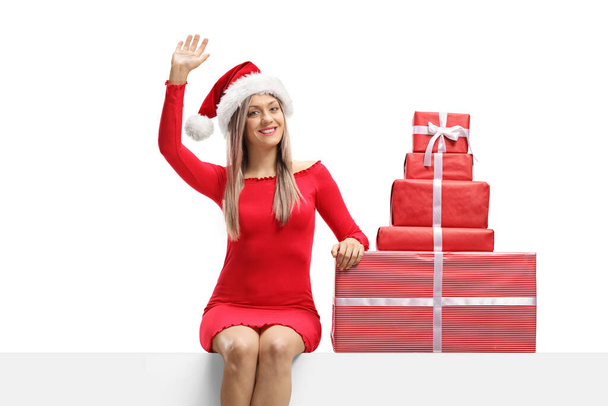 Attractive young female in a red dress and a santa claus hat sitting on a panel with presents and waving isolated on white background - Photo, Image