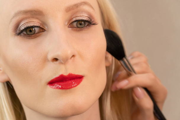 Detailní záběr krásné mladé blondýny s večerní make-up s červenou rtěnkou na rtech s make-up umělce rukou s pudrem štětcem. Soustřeď se na rty - Fotografie, Obrázek