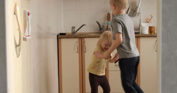 Children dance and jump in the kitchen. Brother and sister have fun together. Blonde sbillings rejoice - Πλάνα, βίντεο