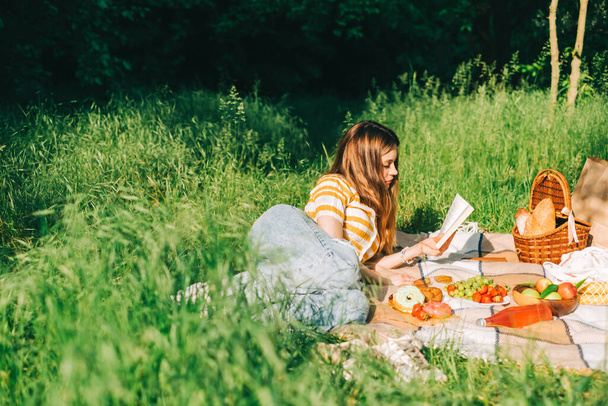 Young woman reading book outdoors, in natural park on picnic, lying on a picnic blanket and enjoying summer weather. - Foto, imagen