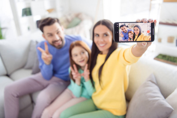 Photo of young attractive cheerful family happy positive smile make selfie smartphone show peace cool v-sign sit couch indoors - Foto, Imagem