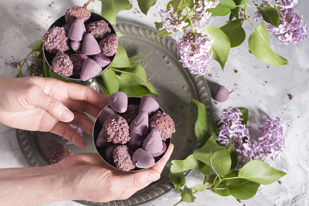 Belgische Bonbons Cuberdon und Pralinen in Silbergläsern, Frühlingsstillleben mit lila Blüten, flache Lage mit weiblichen Händen - Foto, Bild