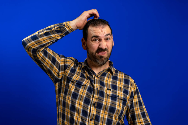 Een man met een baard in een geel geruit hemd die zijn hoofd krabt met een twijfelachtig gebaar geïsoleerd op een blauwe studio achtergrond. Raadsel concept - Foto, afbeelding