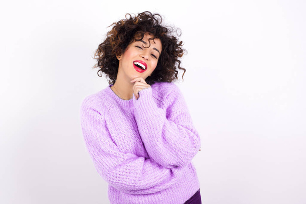  jong mooi latino vrouw dragen trui lacht gelukkig houdt hand op kin uitdrukt positieve emoties glimlacht breed heeft zorgeloze uitdrukking - Foto, afbeelding