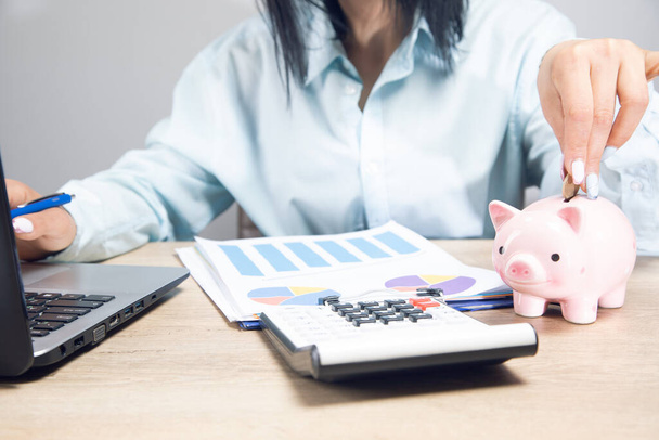 μια γυναίκα κάθεται μπροστά από ένα τραπέζι εργασίας και ρίχνει χρήματα σε ένα κουμπαρά σε ένα γκρι φόντο - Φωτογραφία, εικόνα
