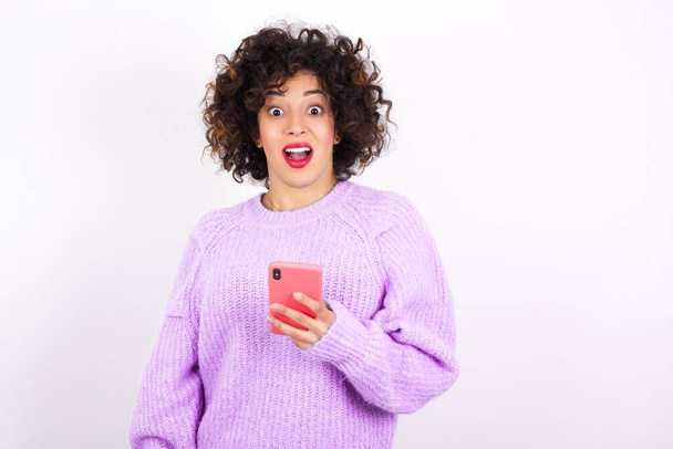young beautiful latin woman wearing sweater  holds smartphone in hands and rejoices positive news, uses modern phone - Foto, Bild