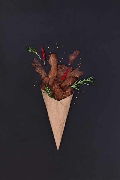 Jerky snacks, red papper and rosemary in craft paper bag on black background. Dried and spiced meat for beer. - Fotó, kép