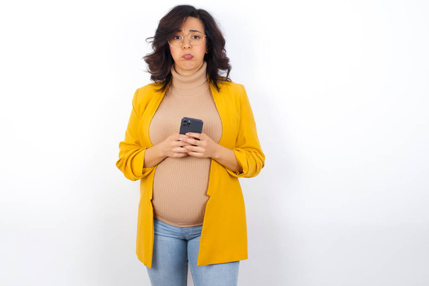 Portrait of a confused pregnant woman holding mobile phone and shrugging shoulders and frowning face. - Foto, afbeelding