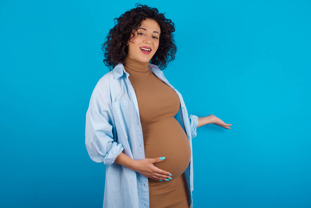  έγκυος γυναίκα Άραβας αισθάνεται ευτυχισμένη και χαρούμενη, χαμογελώντας και καλωσορίζοντάς σας, προσκαλώντας σας σε με μια φιλική χειρονομία - Φωτογραφία, εικόνα