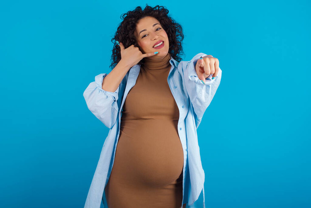  mujer árabe embarazada sonriendo alegremente y apuntando a la cámara mientras hace una llamada más tarde gesto, hablando por teléfono - Foto, imagen