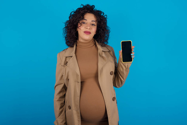 Улыбающаяся беременная женщина держит мобильный телефон с пустым пустым экраном - Фото, изображение