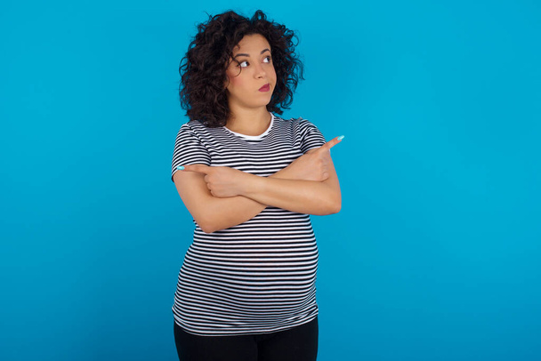  pregnant arab woman wearing striped T-shirt crosses arms and points at different sides hesitates between two items or variants. Needs help with decision - Foto, Imagem