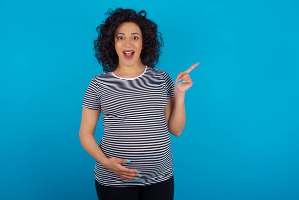  pregnant arab woman wearing striped T-shirt points aside on copy blank space. People promotion and advertising concept - Photo, Image