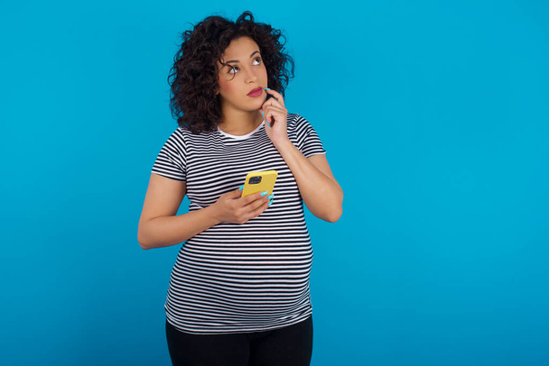 беременная женщина о чем-то глубоко думает, пользуется современным мобильным телефоном, пытается придумать хорошее сообщение, держит указательный палец у губ. - Фото, изображение