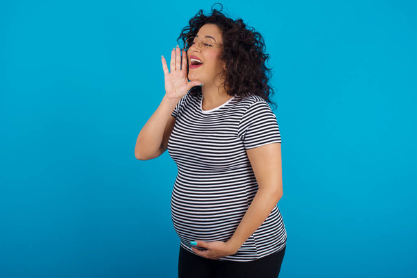  mujer árabe embarazada vistiendo una camiseta a rayas mirando el espacio vacío sosteniendo la mano cerca de su cara y gritando o llamando a alguien. - Foto, Imagen