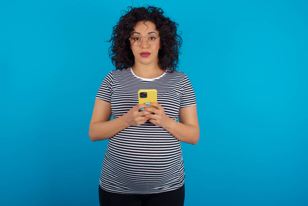 Porträt einer ernsthaften selbstbewussten Schwangeren, die ihr Telefon in zwei Händen hält - Foto, Bild