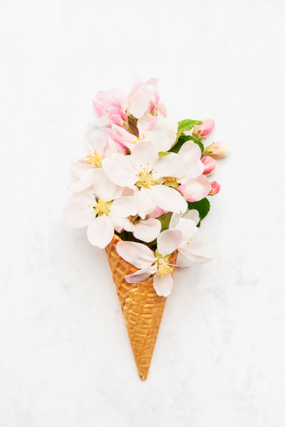 Waffle ice cream cone with apple blossom flowers on a white marble background. Spring concept. Copy space for text, top view - 写真・画像
