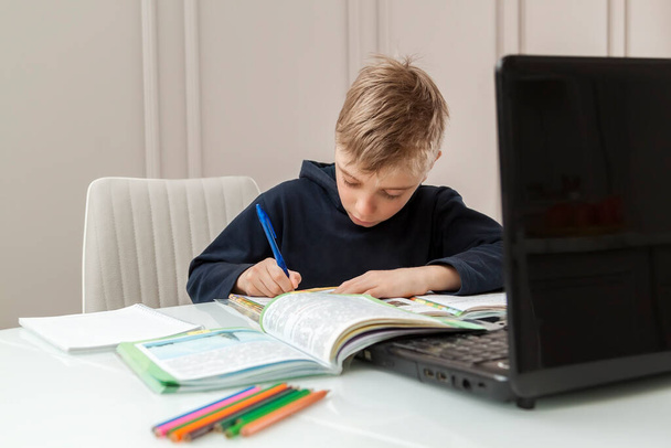 boy writes in notebook tasks received from teacher on laptop screen. Smart Little Boy Is doing His Homework in Living Room. COVID-19 pandemic forces children online learnin - Zdjęcie, obraz